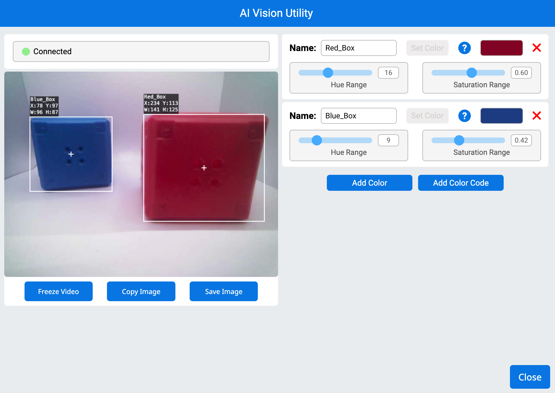 AI Vision Utility muestra un sensor de visión conectado que detecta dos objetos de colores. El lado izquierdo muestra una transmisión de cámara en vivo con un cuadro azul a la izquierda y un cuadro rojo a la derecha, cada uno delineado con cuadros delimitadores blancos. Las etiquetas negras muestran sus respectivos nombres, coordenadas y dimensiones. El lado derecho contiene la configuración de la firma de color, con controles deslizantes para el tono y el rango de saturación para los cuadros rojo y azul. Los botones para añadir colores, congelar vídeo, copiar y guardar la imagen se encuentran en la parte inferior, junto con un botón de cierre en la esquina inferior derecha.